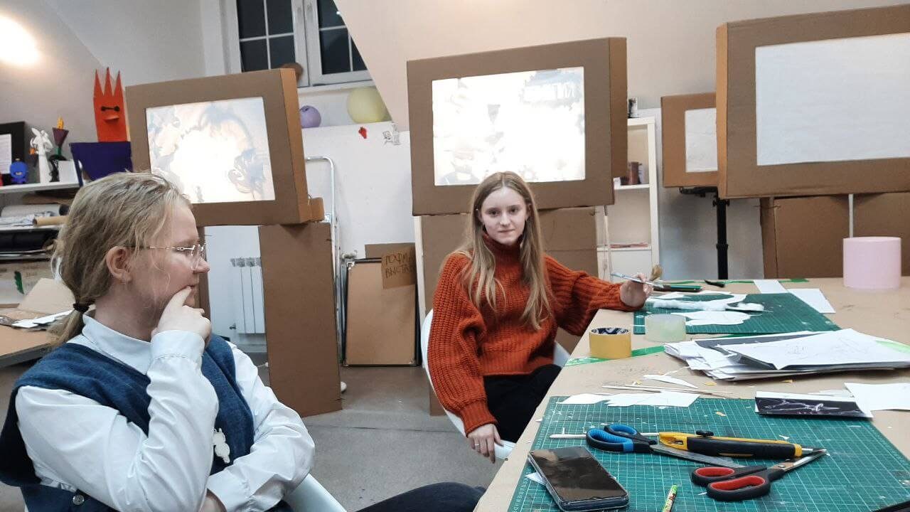 Students cutting silhouettes at a table, with lit shadow theater boxes in the background