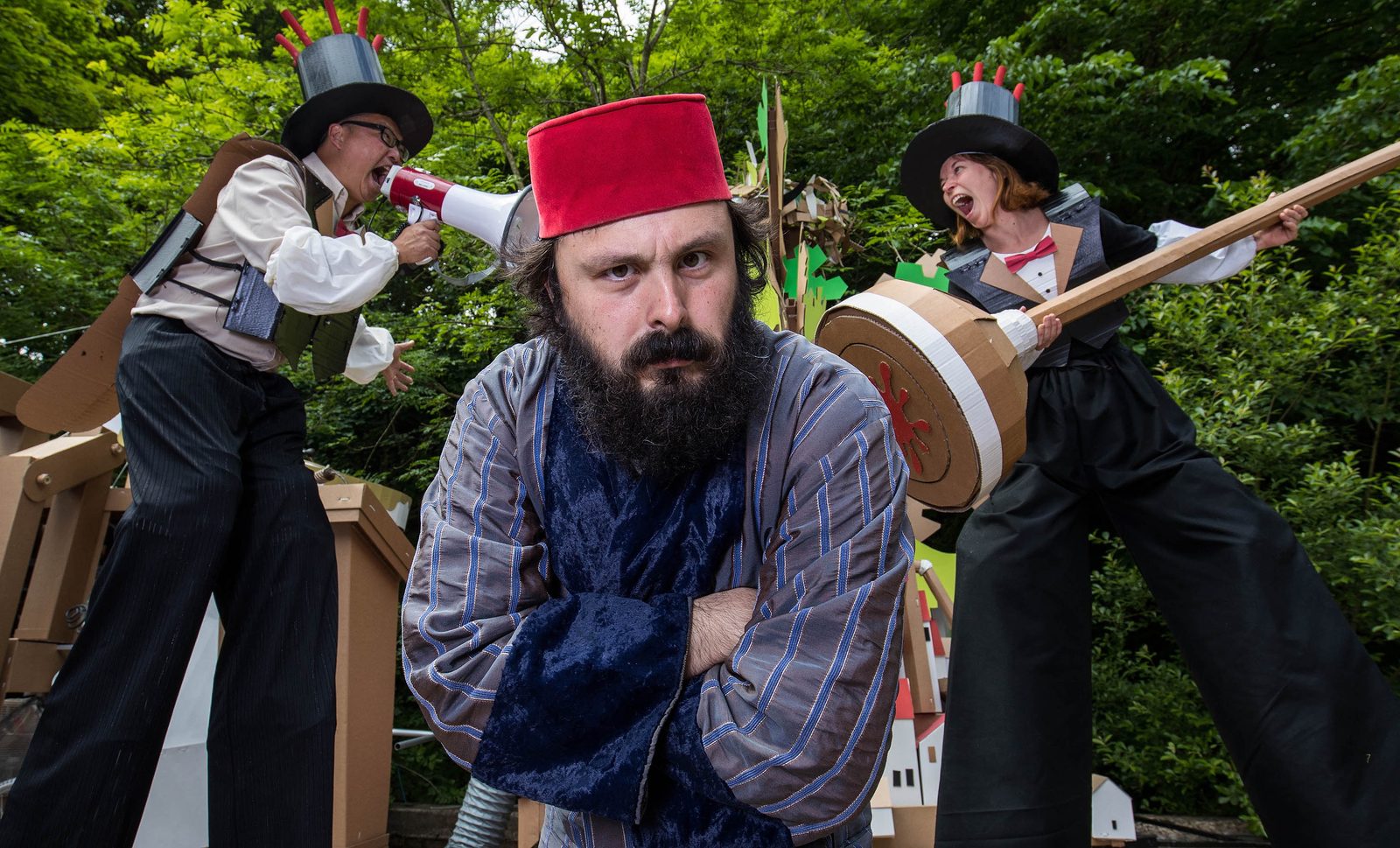 Performers on stilts in cardboard costumes and top hats, one shouting through a megaphone