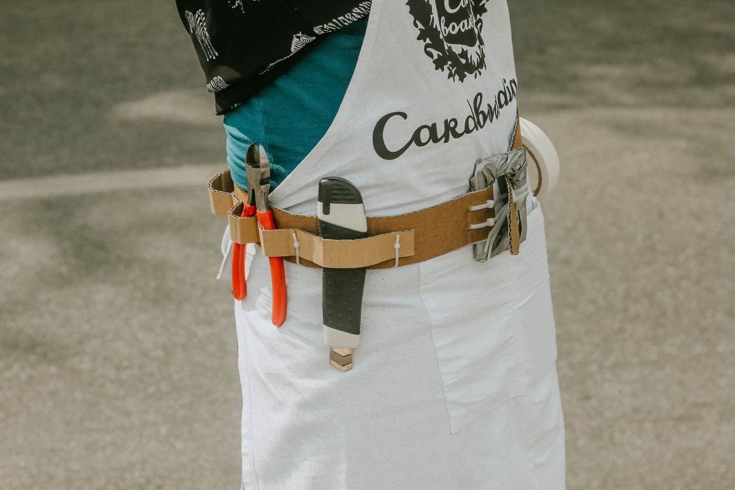 A cardboard tool belt holding real pliers and a box cutter, worn over a Cardboardia apron